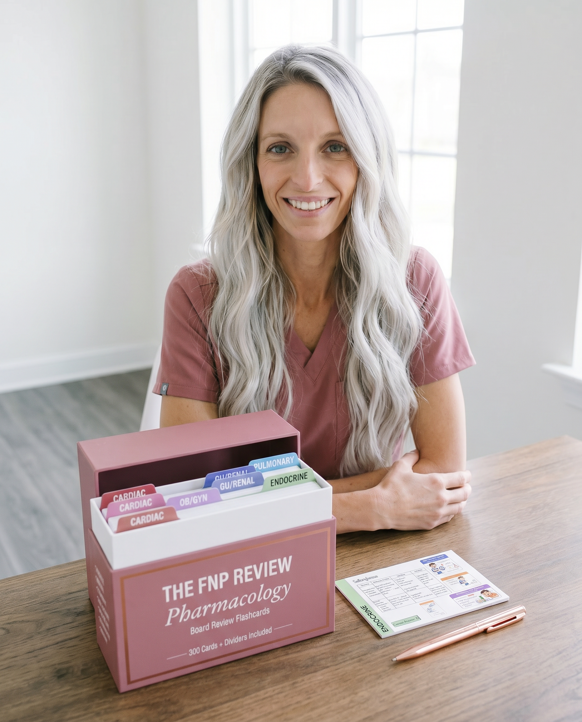 FNP student in pink scrubs smiling with The FNP Review Pharmacology Board Review Flashcard box on desk — 300 cards with dividers